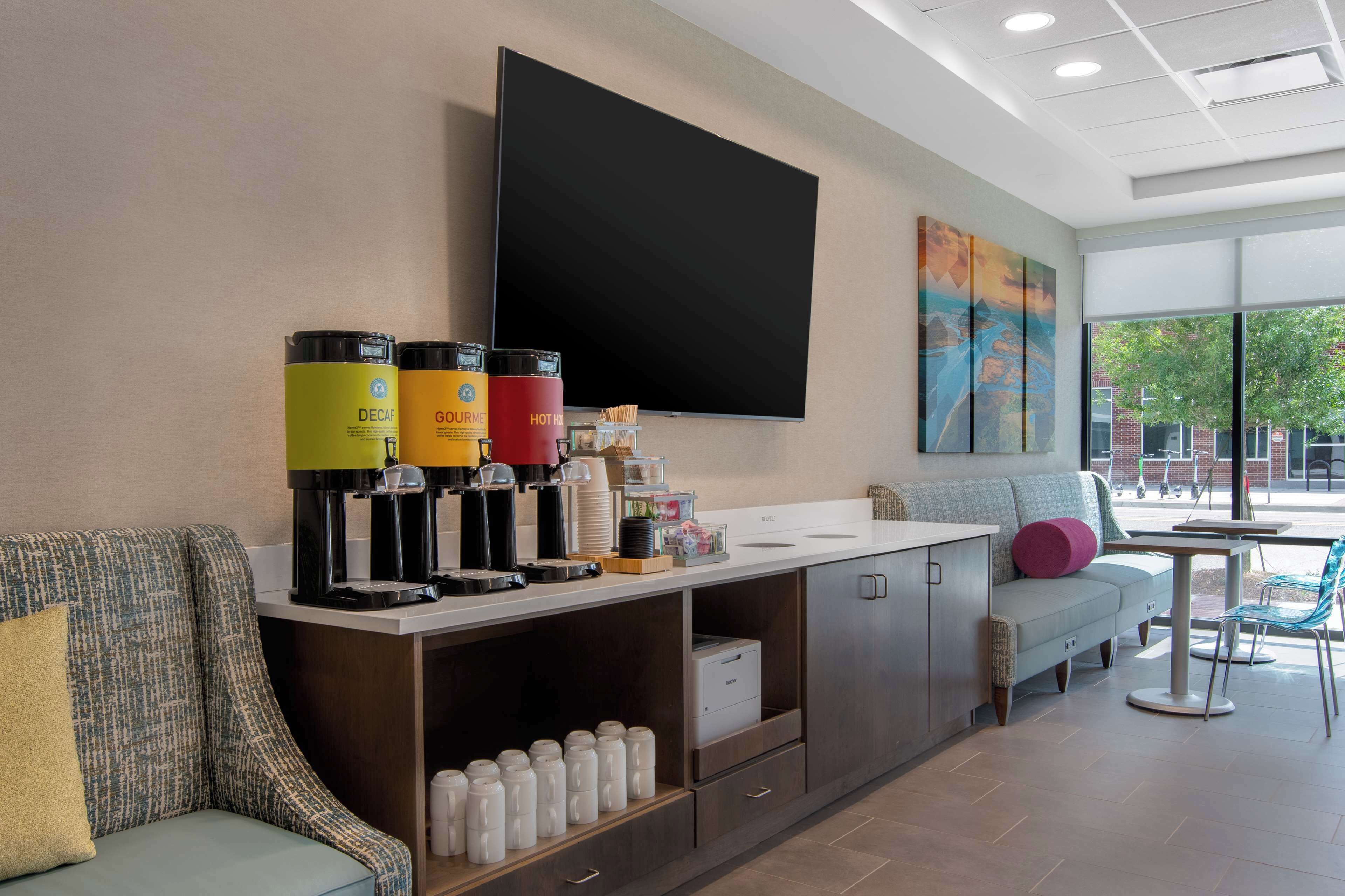 Un moderno hall de hotel con un sofá, una estación de café y un televisor de pantalla plana montado en la pared.