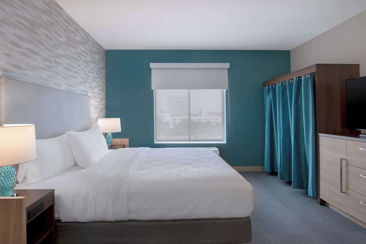 A hotel room with a large bed, a television, a desk with a lamp, a window with curtains, and a view of buildings outside.