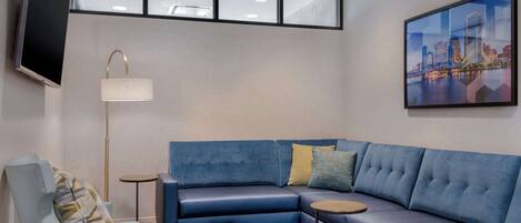 Una habitación de hotel moderna con un sofá seccional azul, una mesita redonda pequeña, una lámpara de pie y un ventanal con vista a la ciudad.