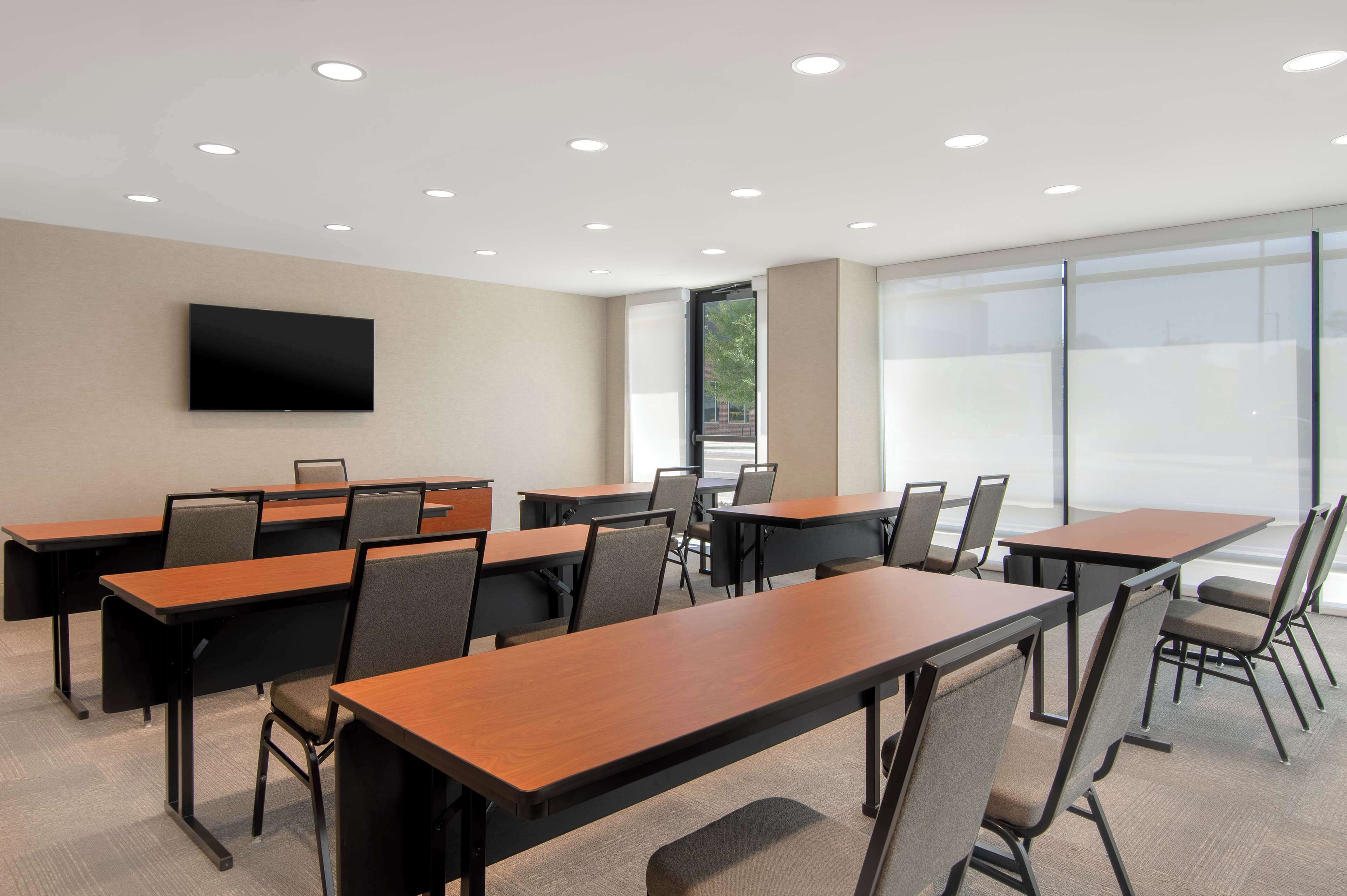 A conference room with multiple tables and chairs, a flat-screen TV mounted on the wall, and large windows providing natural light.