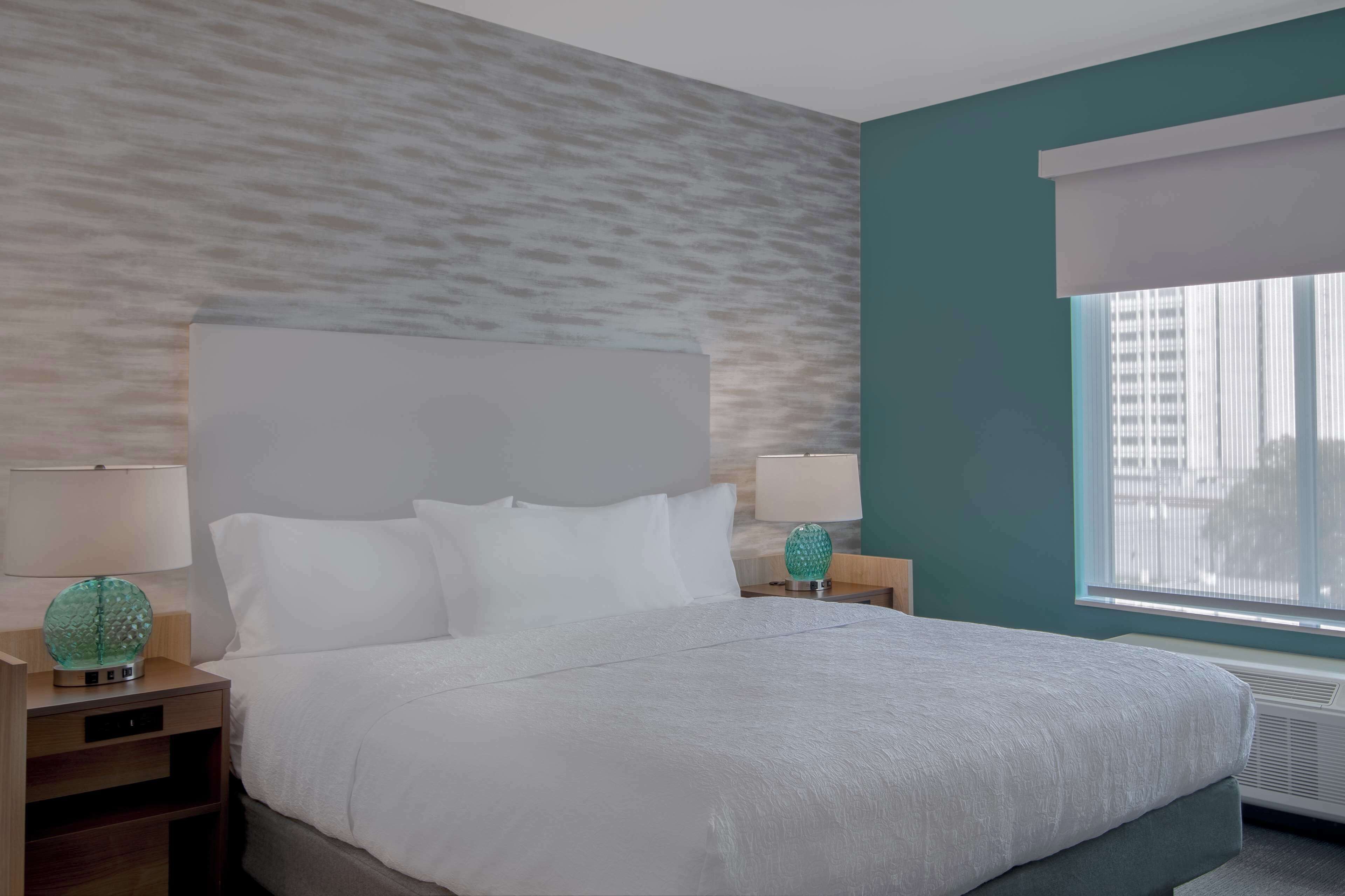 A neatly made bed in a hotel room with a textured wall, a window with blinds, and two bedside tables with lamps.