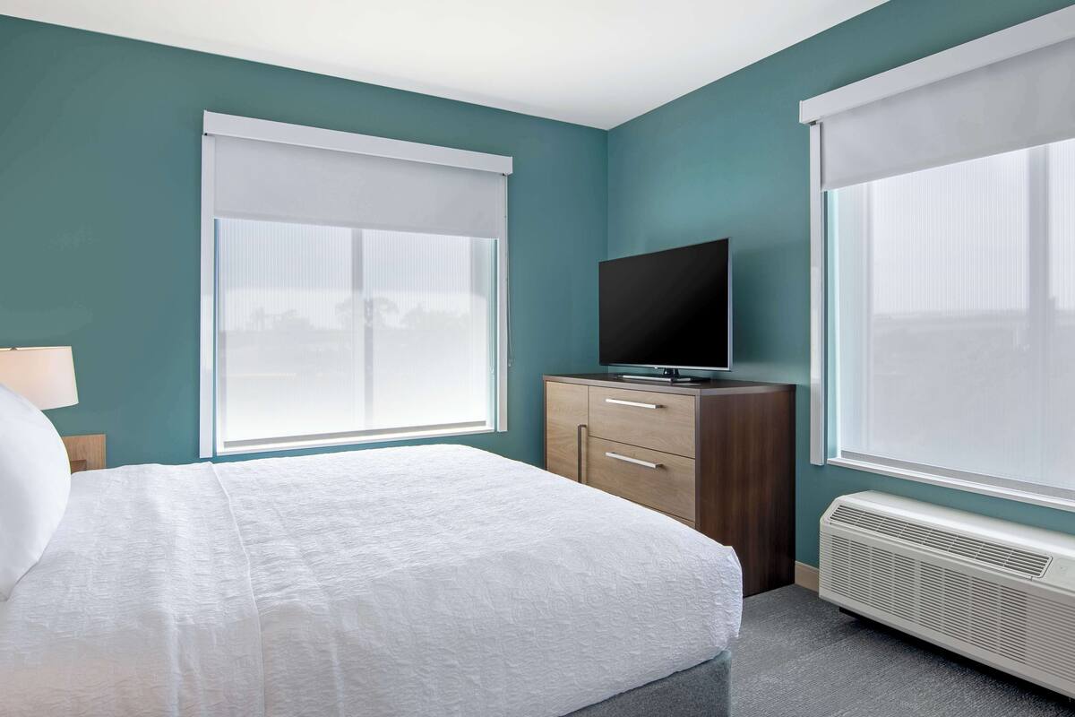 A bedroom with a bed, a TV on a wooden dresser, and a window with a white blind.