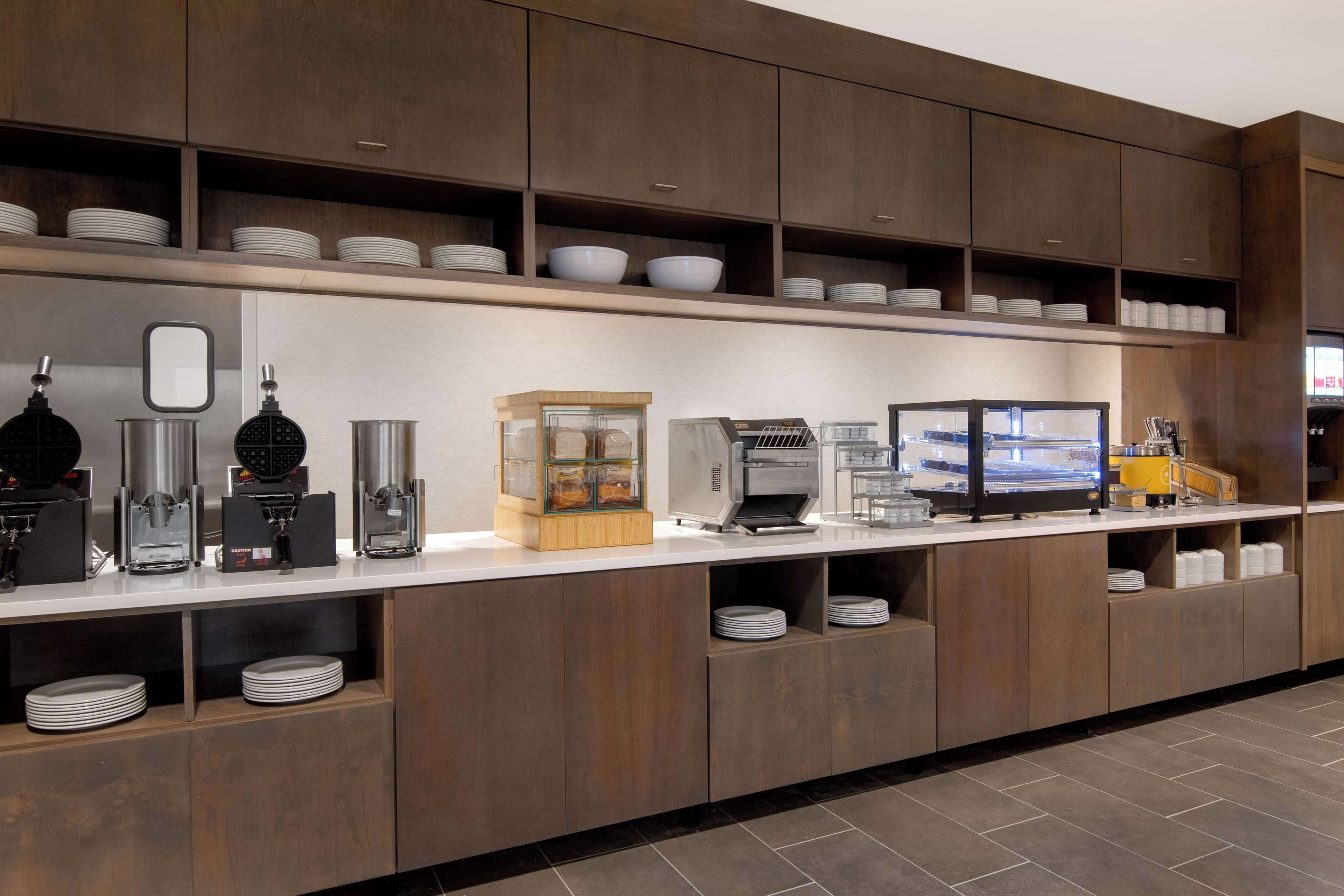 A modern kitchen with wooden cabinets, a white countertop, and various appliances like coffee makers and a food display unit.