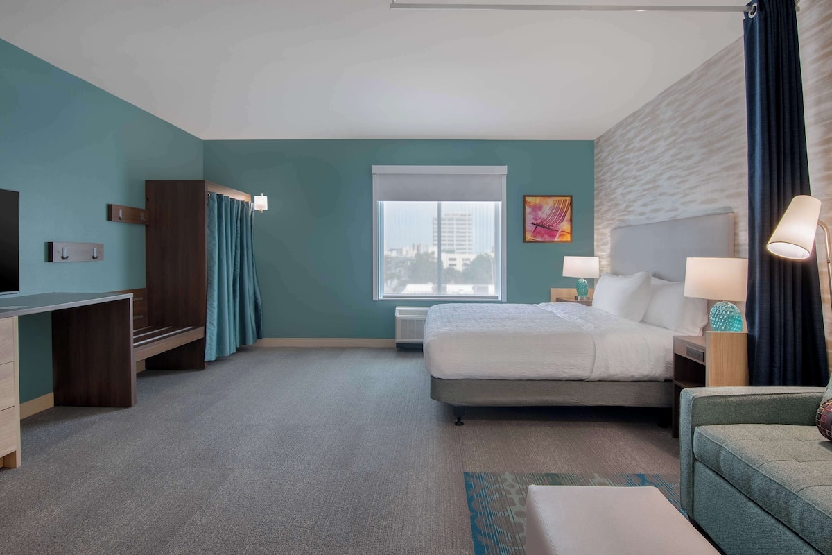 A hotel room with a bed, a desk, a chair, a TV, a window, and a view of the city.
