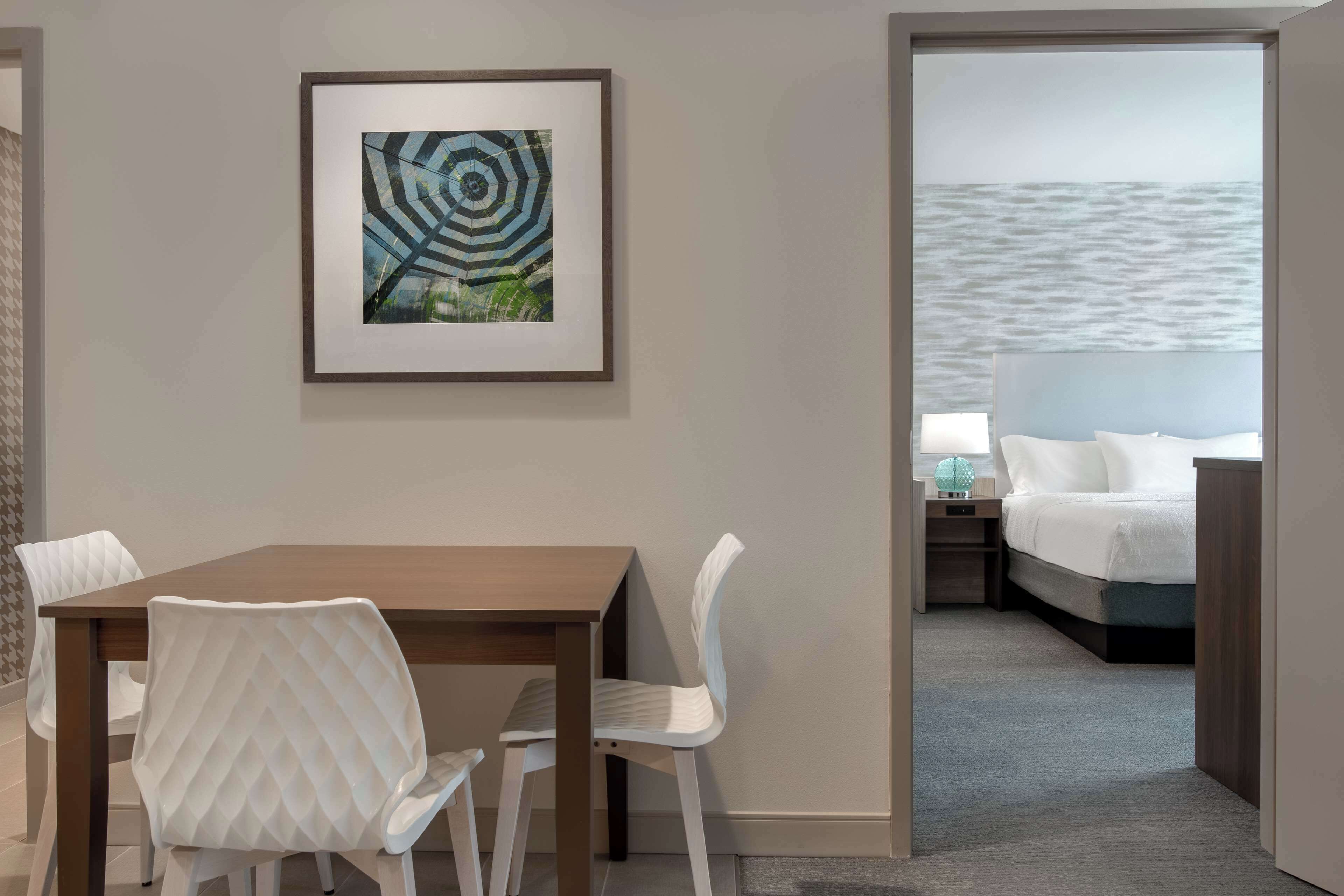 A hotel room with a dining area, a bed with a view of the sea, and a framed artwork on the wall.