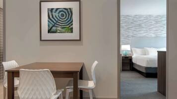 A hotel room with a dining area, a bed with a view of the sea, and a framed artwork on the wall.
