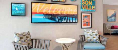 A lobby with two wooden chairs with patterned cushions, a round white table, and a large welcome sign.