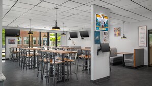A modern indoor dining area with multiple tables and chairs, a high ceiling with grid-like patterns, and various wall decorations including sports team logos.