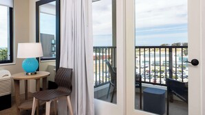 Un balcón con vista a un estacionamiento y edificios, una silla, una mesita con una lámpara y una ventana con cortina.