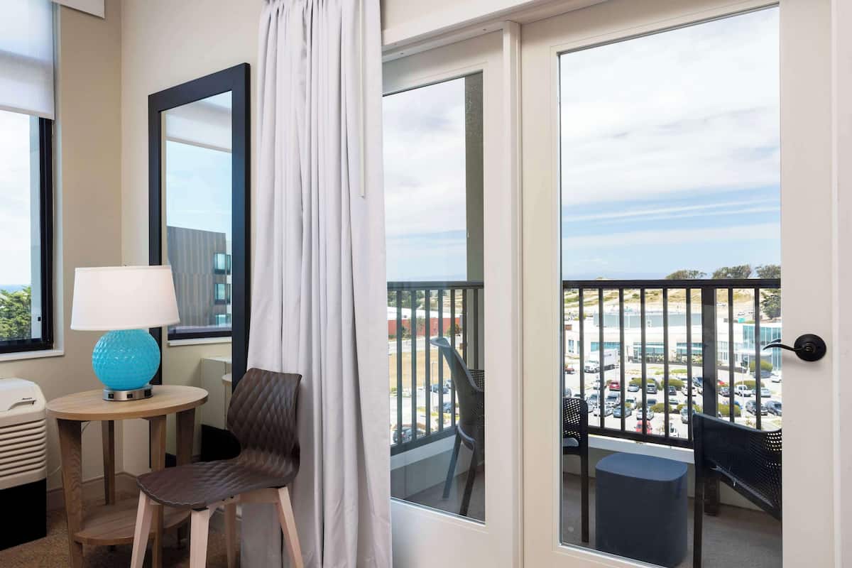 Un balcón con vista a un estacionamiento y edificios, una silla, una mesita con una lámpara y una ventana con cortina.