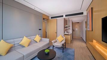 A modern hotel room with a grey sofa, a small round table, a white armchair, and a TV on the wall.
