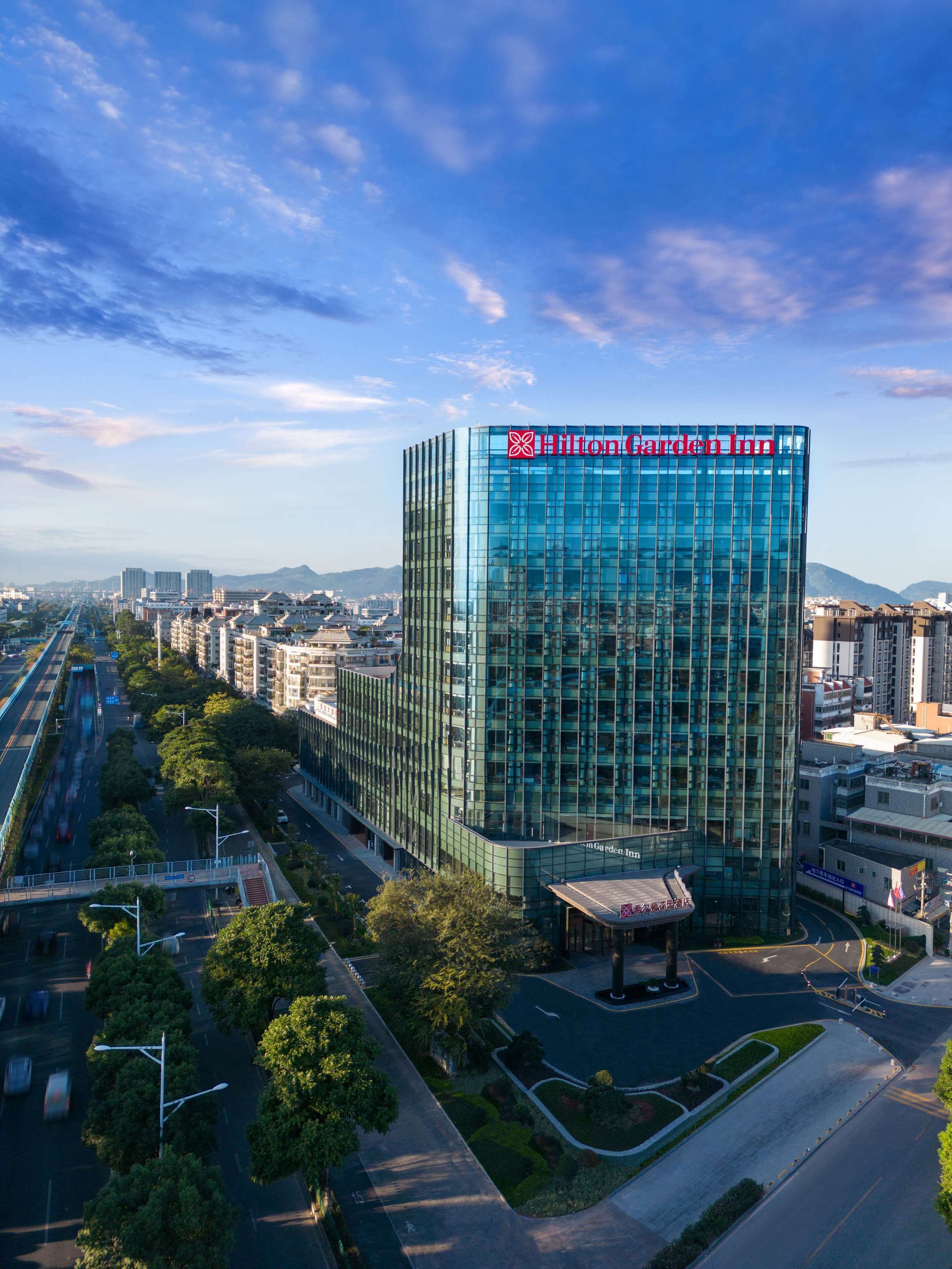 A modern Hilton Garden Inn hotel with a glass facade, surrounded by urban infrastructure and greenery.