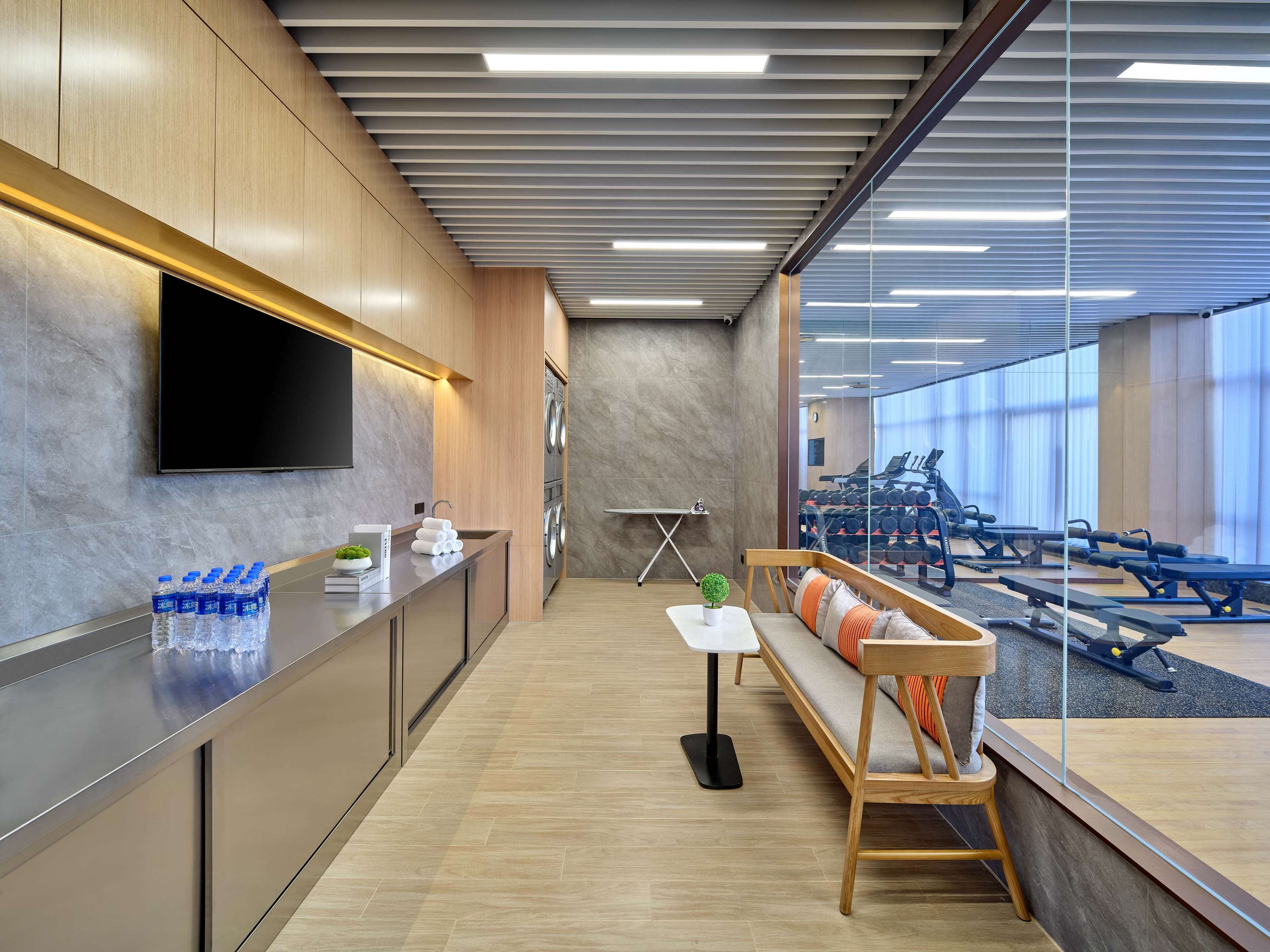Un hall d’hôtel moderne doté d’un téléviseur à écran plat, d’un comptoir de réception, d’un espace salon avec un banc et une petite table, et offrant une vue sur la salle de sport grâce à un mur en verre.