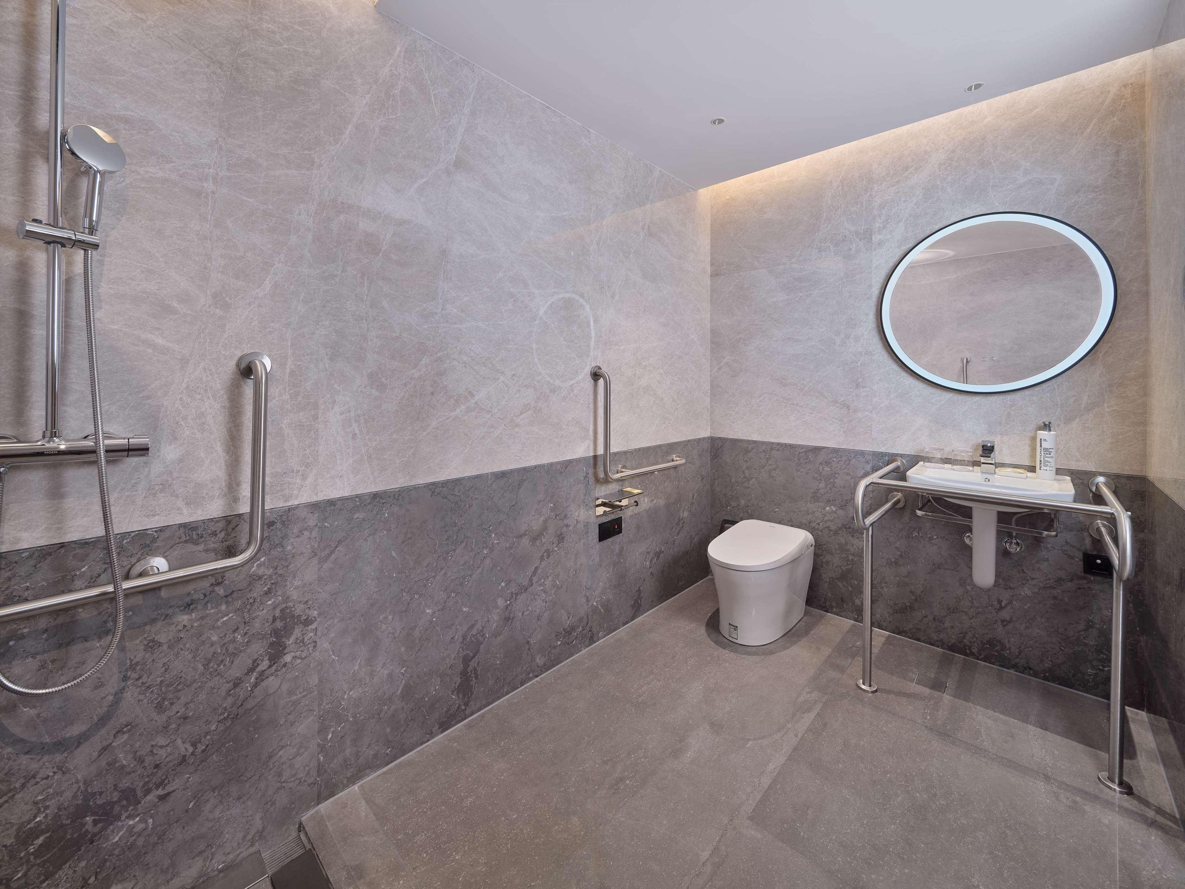 Un baño moderno con piso de mármol, inodoro blanco, lavamanos con espejo y ducha con barras de apoyo.