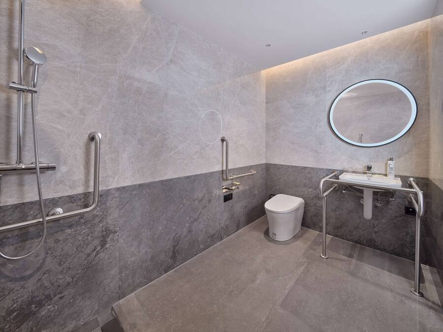 Casa de banho moderna com chão de mármore, sanita branca, lavatório com espelho e chuveiro com barras de apoio.