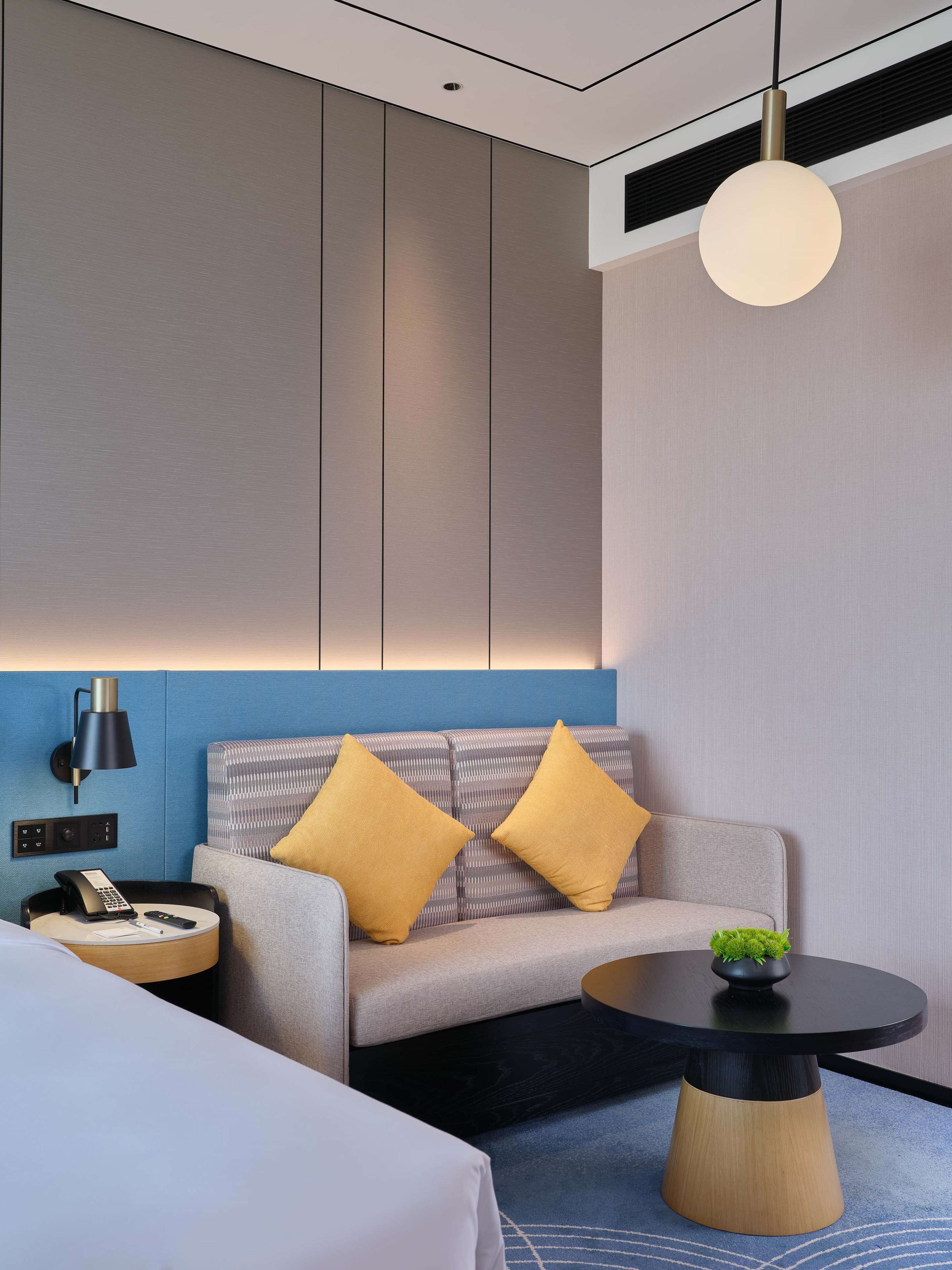 A modern hotel room with a sofa, a bedside table with a lamp, a round coffee table, and a small plant.