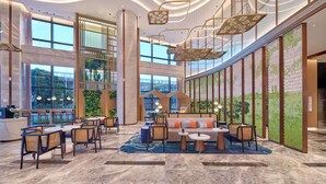 Een ruime lobby met een hoge plafond, moderne verlichtingsarmaturen, een zithoek met banken en tafels, en grote ramen met uitzicht op de omgeving.