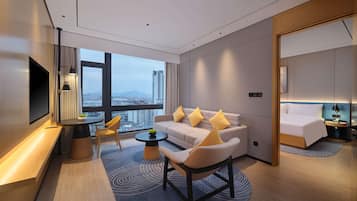 A modern hotel room with a large window, a sofa with yellow cushions, a small round table, and a TV on the wall.