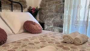 Een net opgemaakt bed met twee opgerolde handdoeken, waarvan er één het woord 'Stella' op heeft staan, in een kamer met stenen muren en een raam met dunne gordijnen.