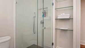 A glass-enclosed shower with a glass door, a white toilet, and a glass shelf with folded towels.
