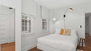 A bedroom with a white bed, a wooden floor, and a single window with curtains.