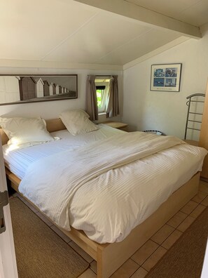 Une chambre avec un grand lit, une table de chevet, une fenêtre avec des rideaux, un tableau encadré au mur et un cadre de lit en métal.