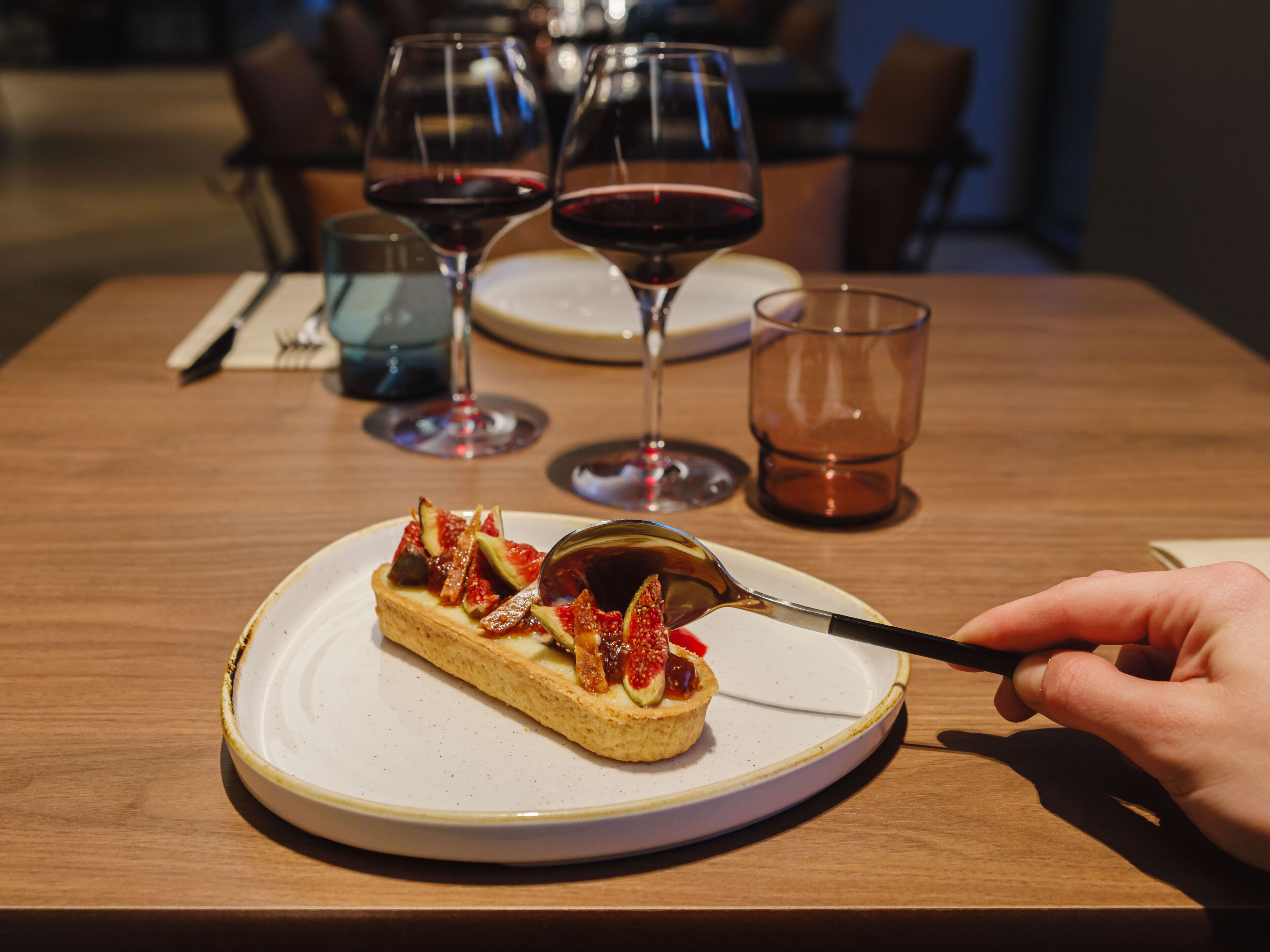 Postre en un plato con cuchara, rodeado de copas de vino y una taza sobre una mesa de madera.