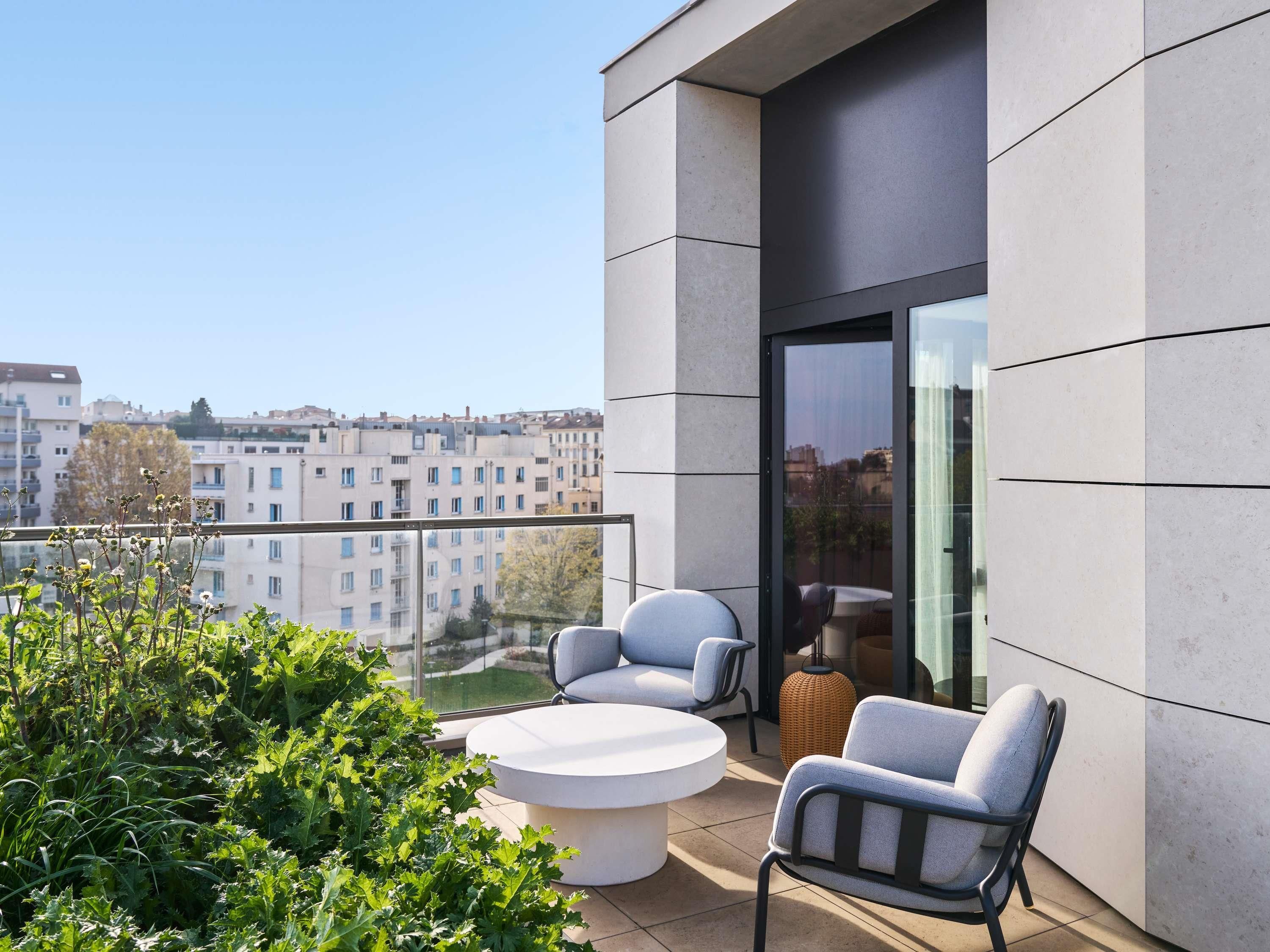 A modern balcony with a round table and two chairs, a wicker ottoman, and a view of the cityscape.