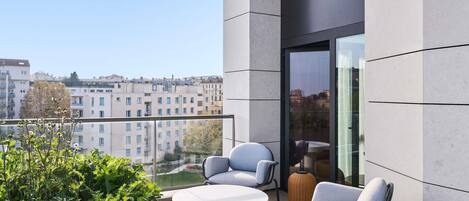 A modern balcony with a round table and two chairs, a wicker ottoman, and a view of the cityscape.