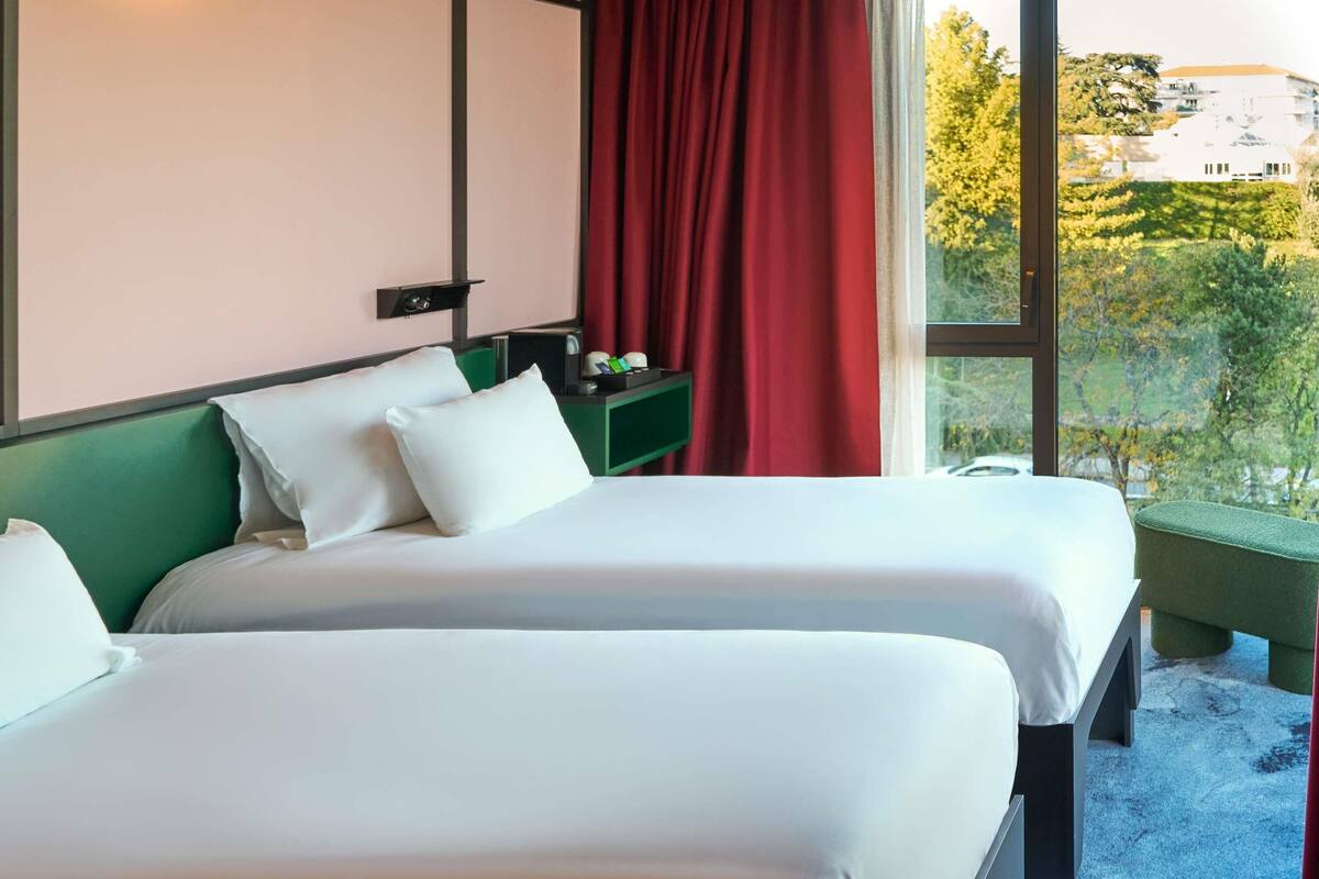 Habitación de hotel con dos camas, cabecera verde, mesita de noche con lámpara y ventana con cortina roja.