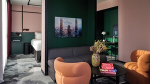 A modern hotel room with a grey sofa, orange armchairs, a small table with a vase and books, a bed with a cityscape painting, and a green accent wall.