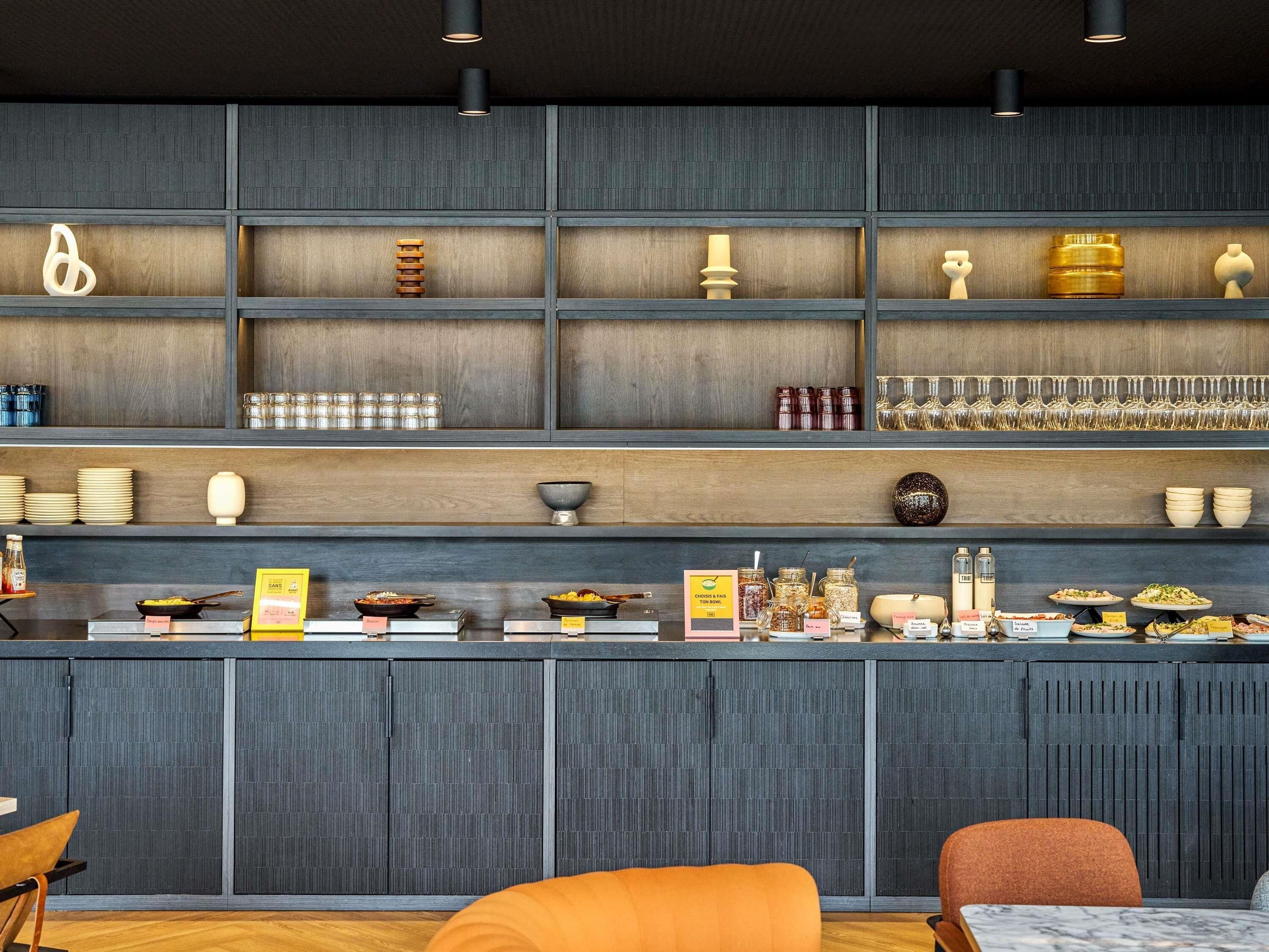 A modern bar area with shelves displaying various glassware and bottles, a counter with food items, and seating with orange cushions.