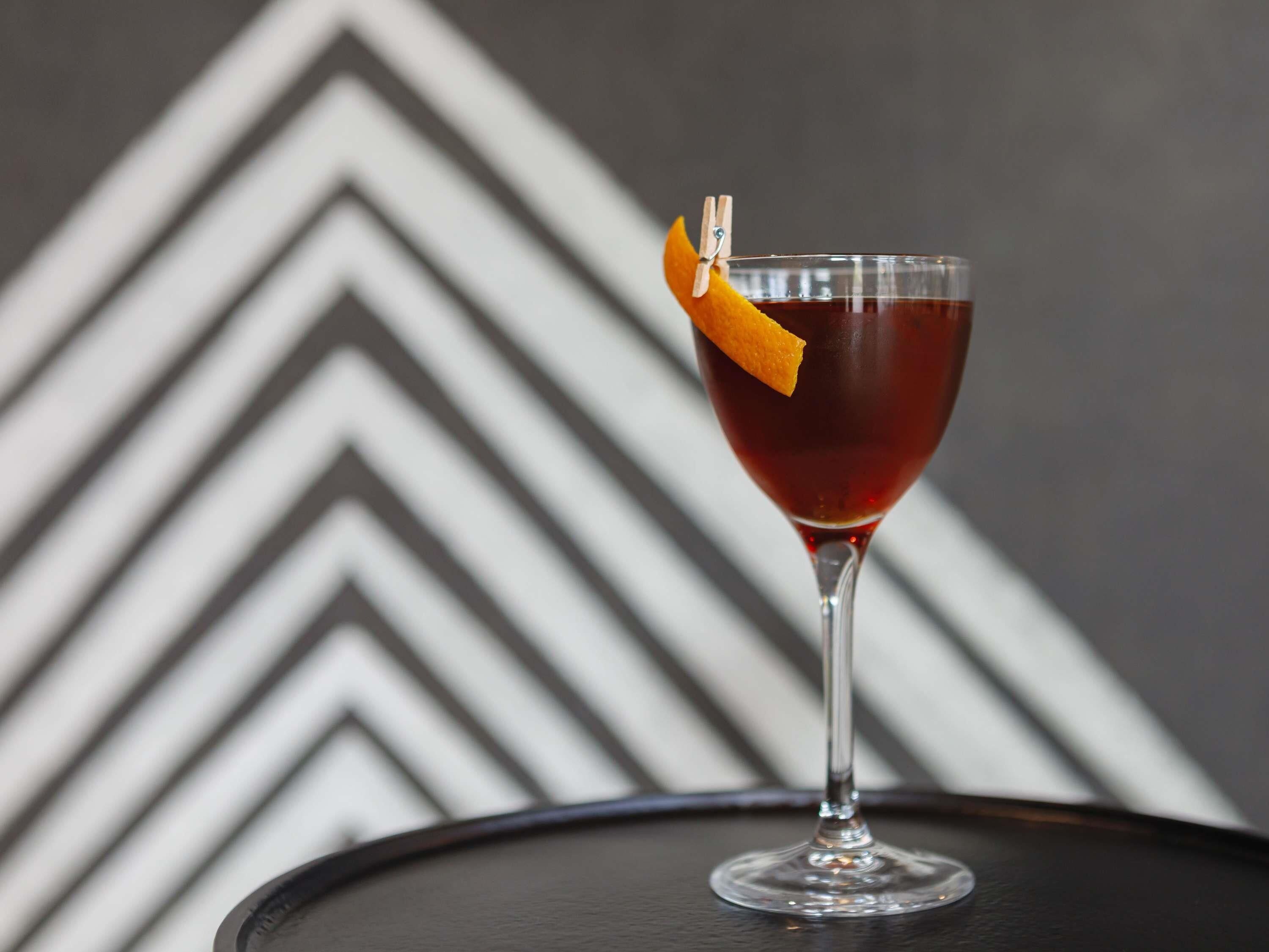 A cocktail in a stemmed glass with an orange peel garnish, set against a geometric patterned background.
