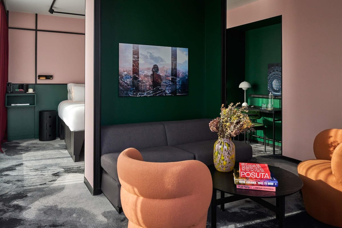 Un hotel moderno con un sofá gris, una mesa de centro con un libro, un jarrón con flores secas y una pintura de un paisaje urbano en la pared.