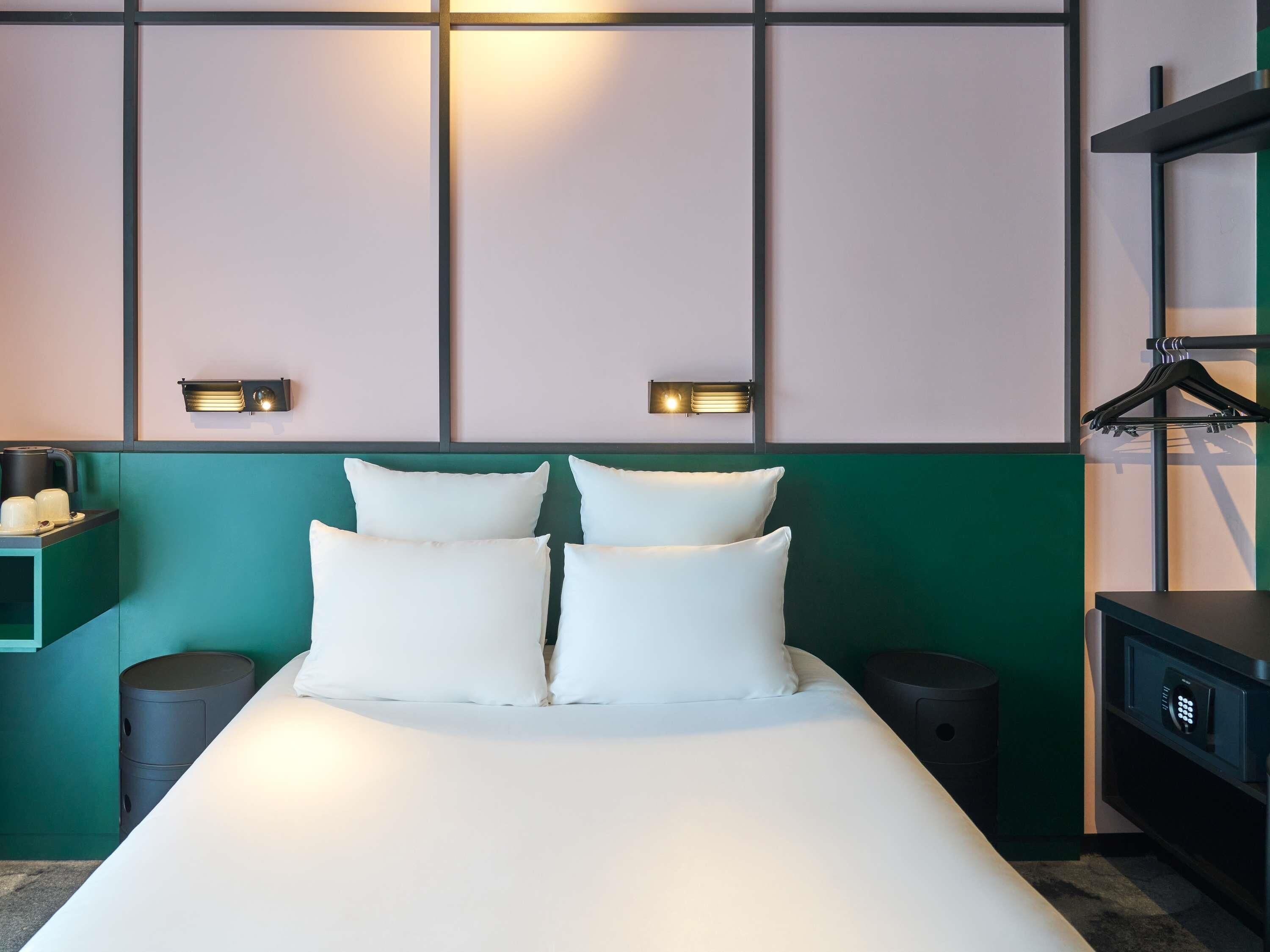 Cama bem arrumada com lençóis e almofadas brancos, mesa de cabeceira com um objeto preto e luminária fixada na parede.