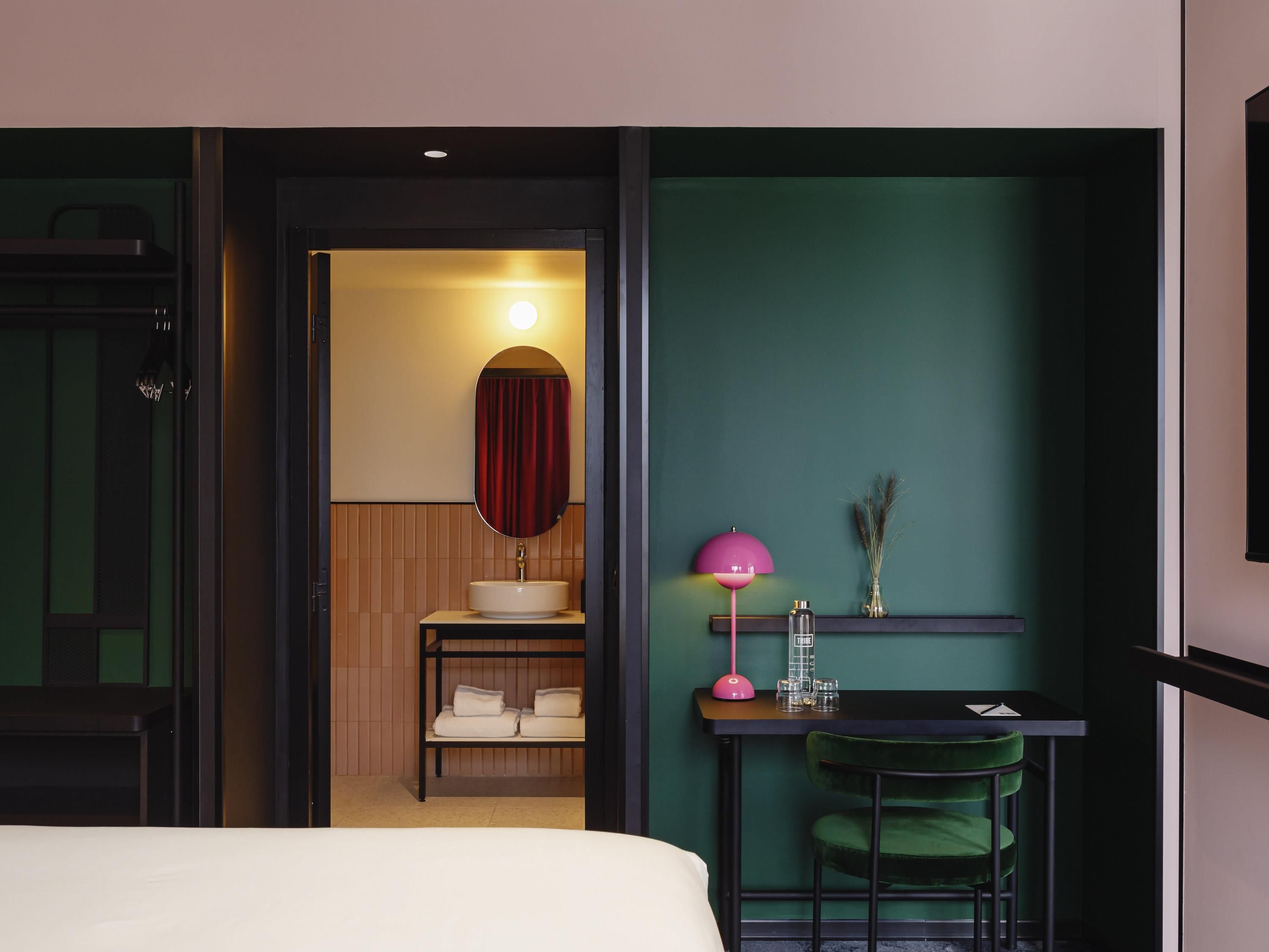 A hotel room with a bed, a desk with a chair, a small table with a lamp, a mirror, and a wall-mounted shelf with decorative items.