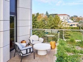 A modern outdoor seating area with a round table and chairs, a wicker lamp, and a view of a cityscape with buildings and trees.