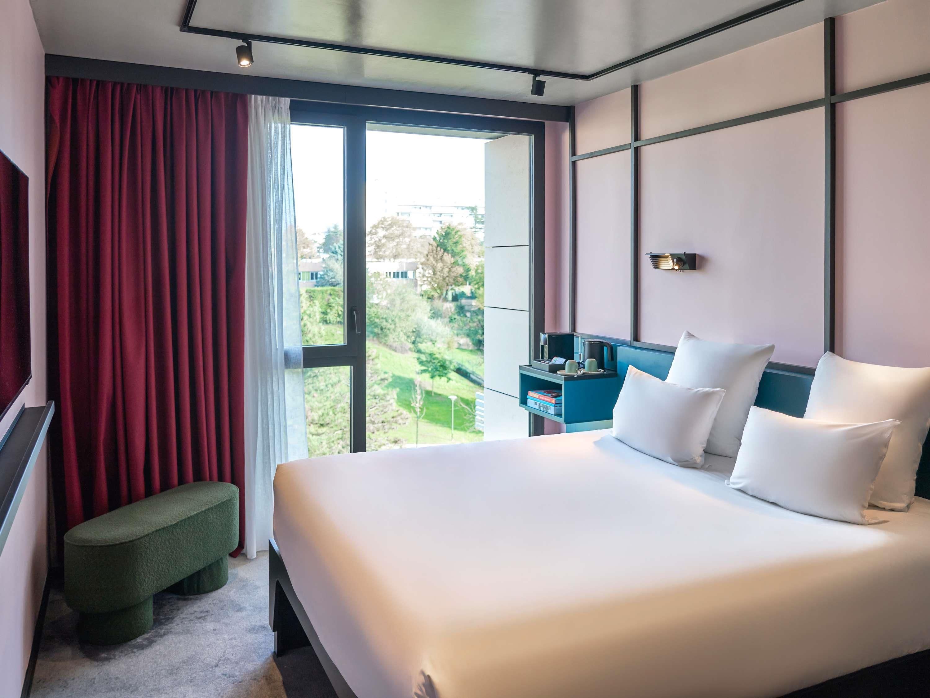 Habitación de hotel con una cama grande, una mesita de noche, un banco y vista a un paisaje verde a través de una ventana.