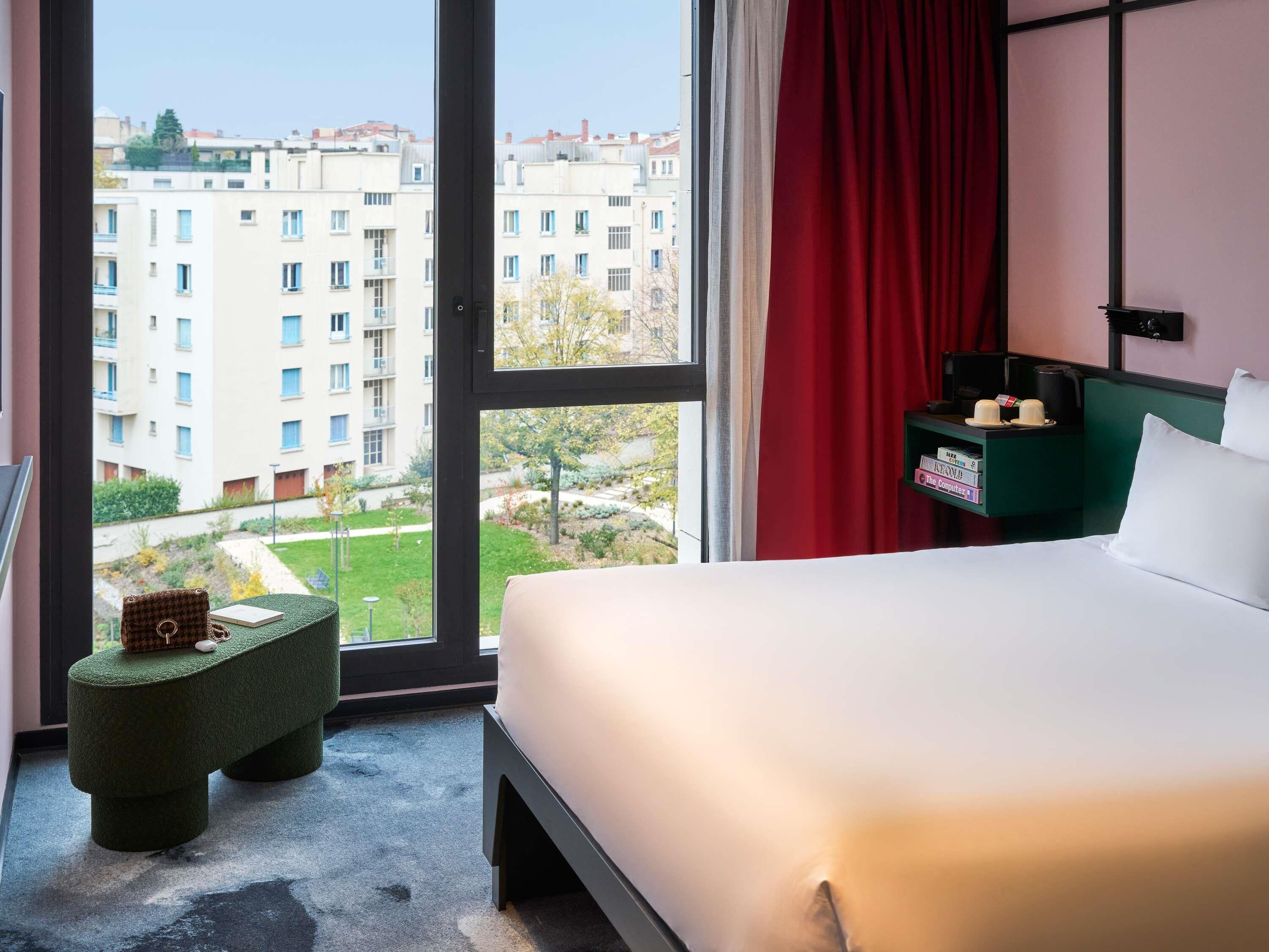 A hotel room with a bed, a small green stool, a nightstand with a lamp, and a view of buildings through a large window.