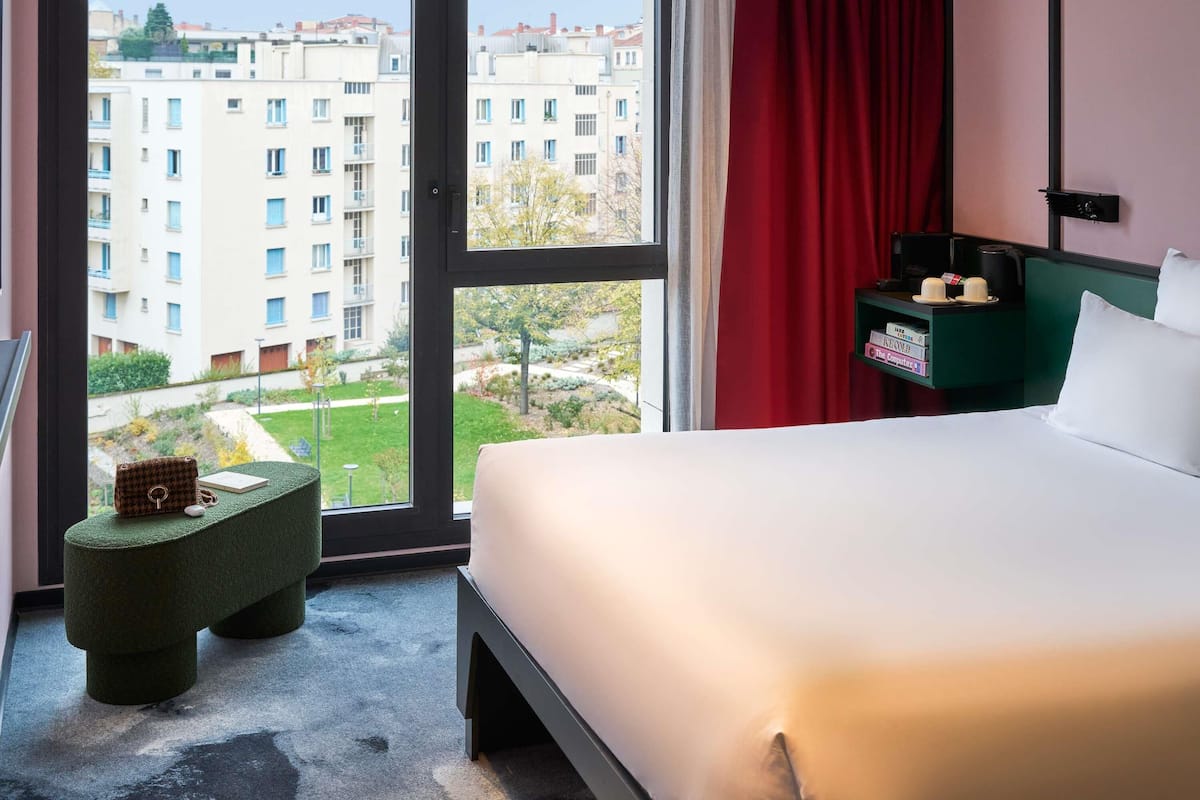 A hotel room with a bed, a small green stool, a nightstand with a lamp, and a view of buildings through a large window.