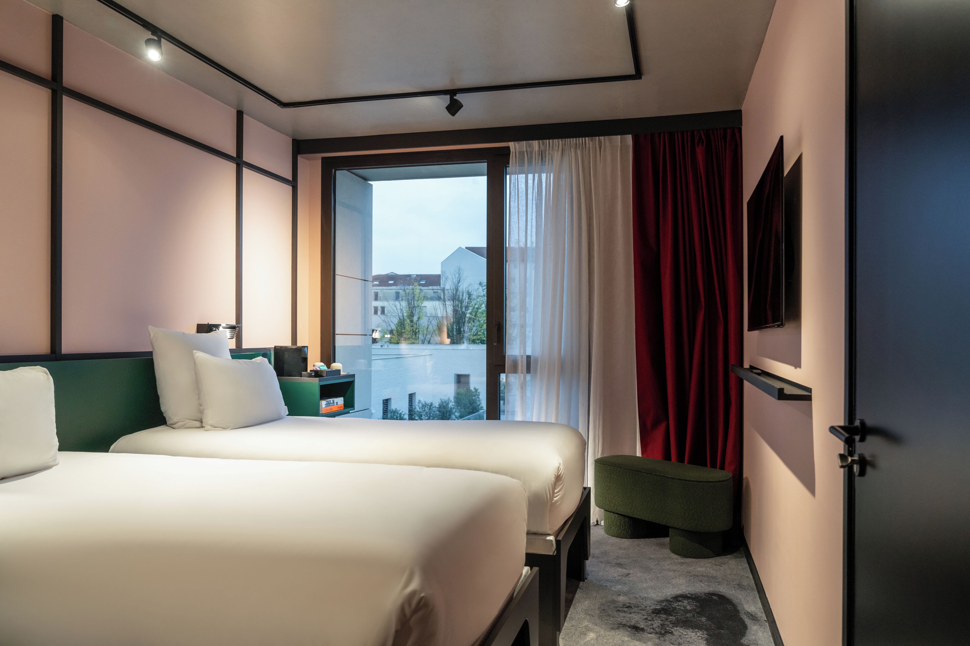 A hotel room with two beds, a green bench, a TV, and a view of buildings through a large window.