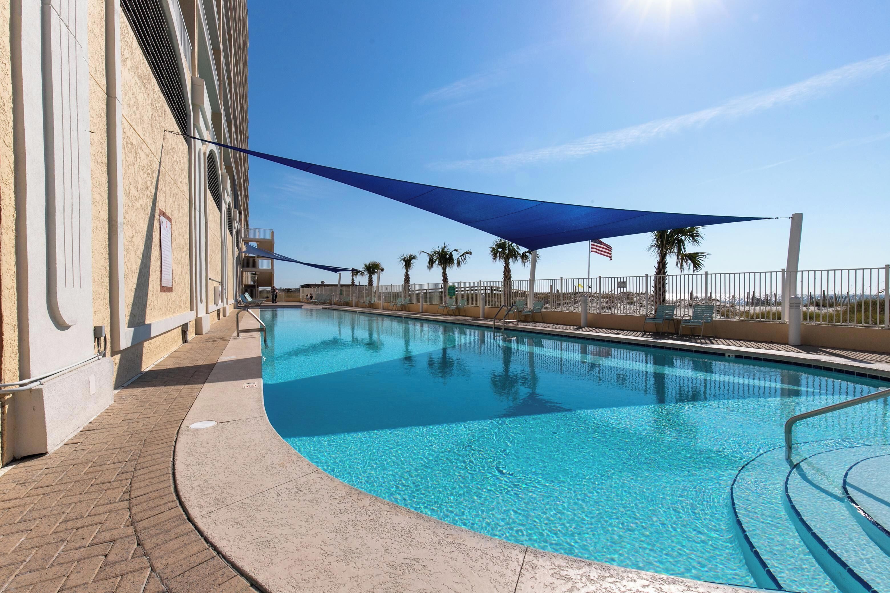 A swimming pool with a clear blue water surface, surrounded by a paved walkway and a shaded area with a blue canopy.