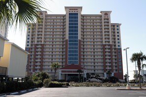A multi-story hotel with a central glass tower, multiple balconies, and a parking lot in front.