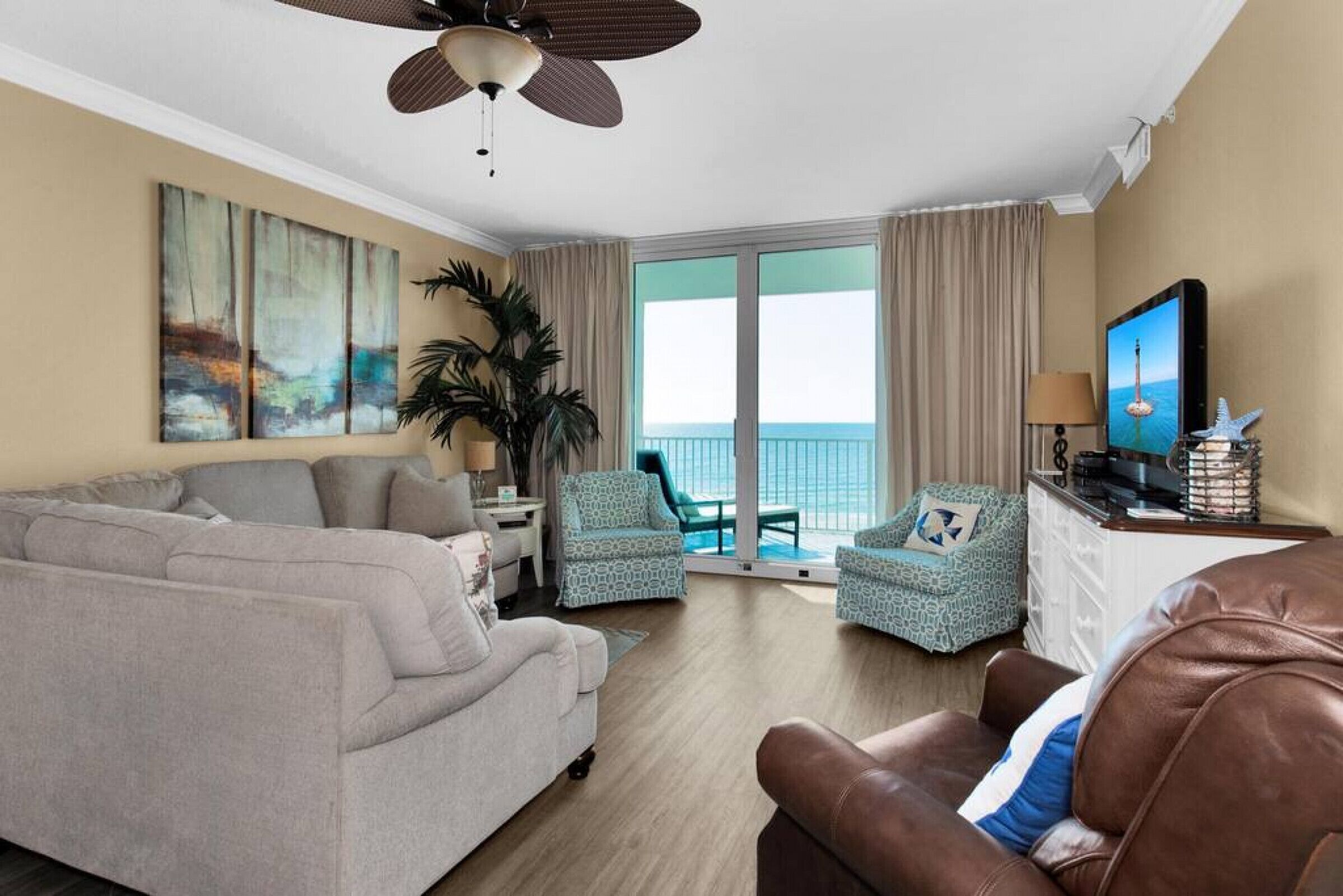 Une chambre d’hôtel avec un canapé, deux fauteuils, une télévision, un ventilateur de plafond, une grande fenêtre avec des rideaux et une plante.
