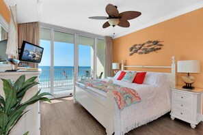A hotel room with a bed, a desk with a computer, a ceiling fan, a balcony with a view of the ocean, and a wall decoration.