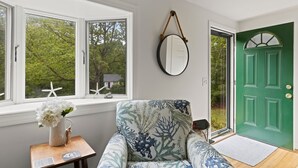 A room with a green door, a white-framed window, a chair with a floral pattern, and a small table with a vase of flowers. - 15 Courtland Drive (Sandwich)