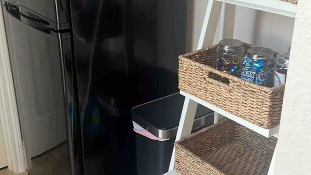 A compact kitchen with a black refrigerator, a trash bin, and a white shelving unit with wicker baskets. - Lake-Themed Private Guest Suite Gated Community Pet Friendly Near I-4 (Plant City)