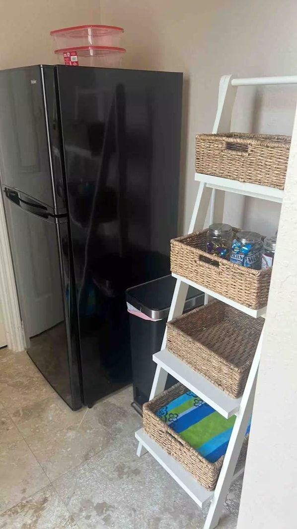 A compact kitchen with a black refrigerator, a trash bin, and a white shelving unit with wicker baskets. - Lake-Themed Private Guest Suite Gated Community Pet Friendly Near I-4 (Plant City)