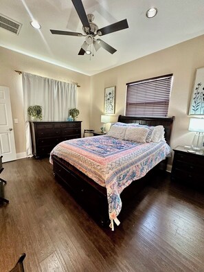 Un dormitorio con cama, ventilador de techo, ventana con persianas, cómoda y mesita de noche.