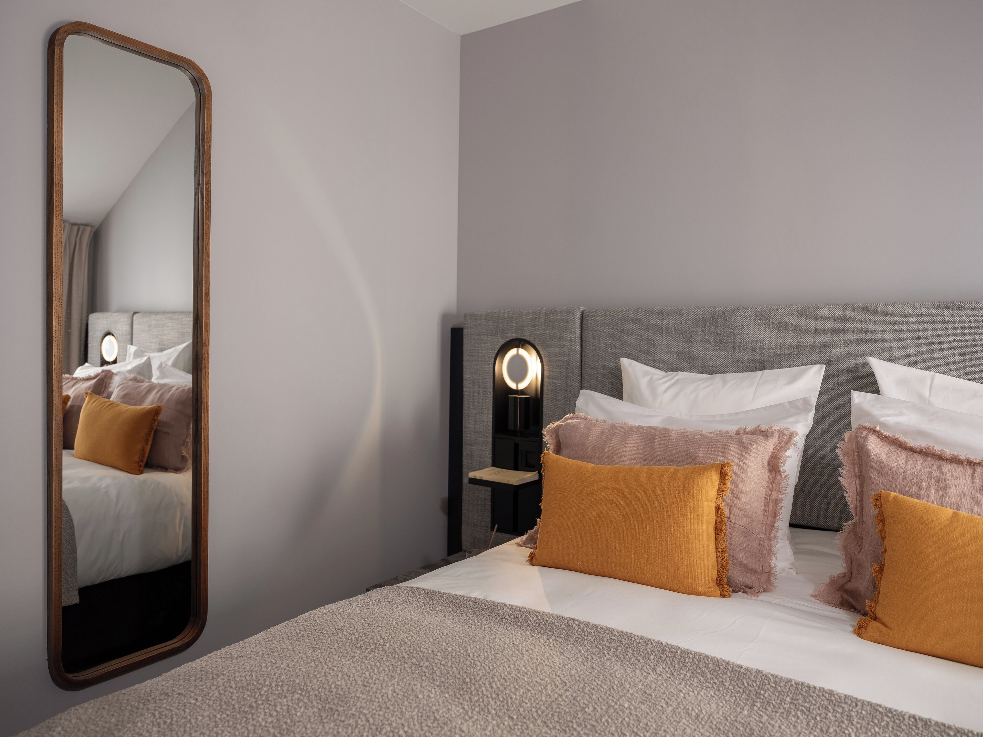 Ein modernes Schlafzimmer mit einem großen Spiegel, einem Bett mit weißen und senffarbenen Kissen, einem Kopfteil und einem Nachttisch mit Lampe.